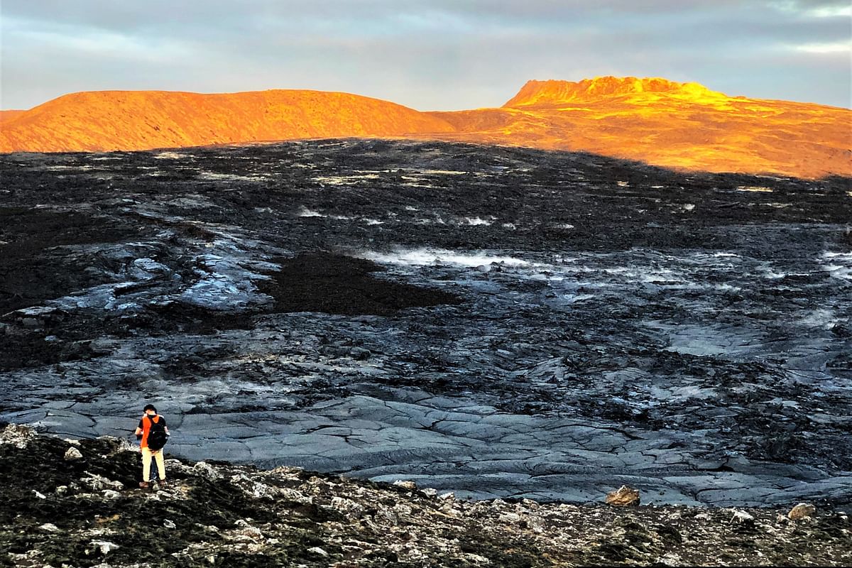 Hiker explores rugged volcanic landscape during sunset with vivid orange glow on distant hills.