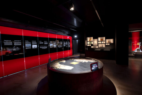 Exhibit room with illuminated displays and interactive table, showcasing historical artifacts and information panels.