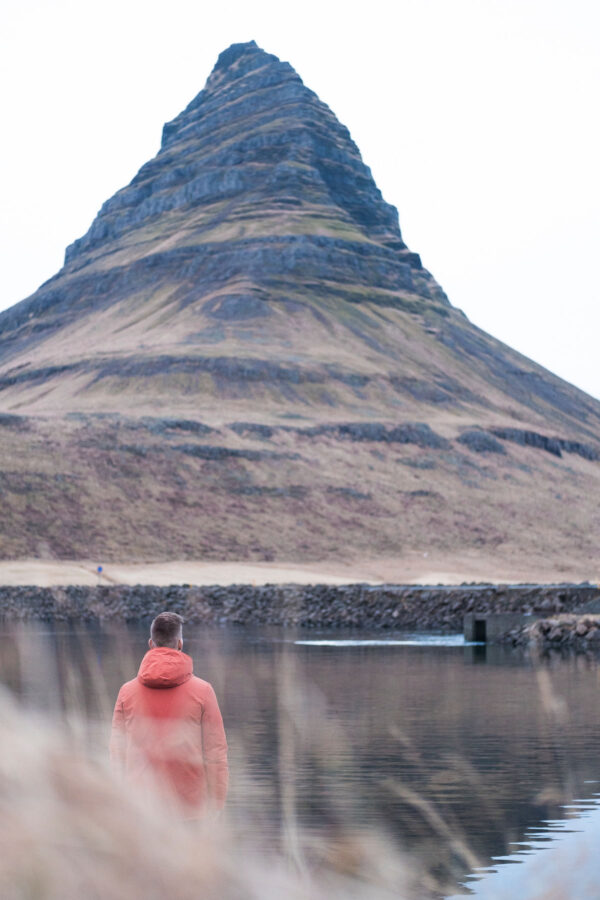 Person in a red jacket stands by a calm lake, gazing at a rugged mountain. Tranquil nature scene.
