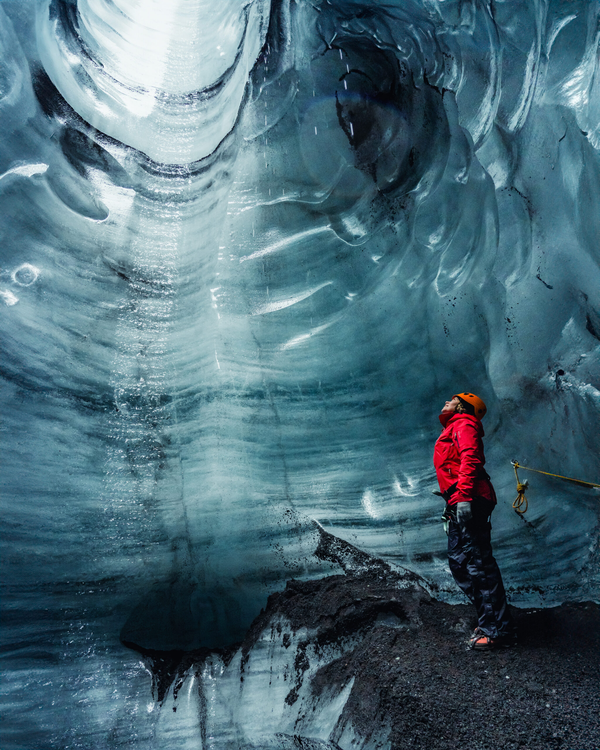 Person in a red jacket and helmet standing inside an icy cavern, looking up at the towering blue ice formations.