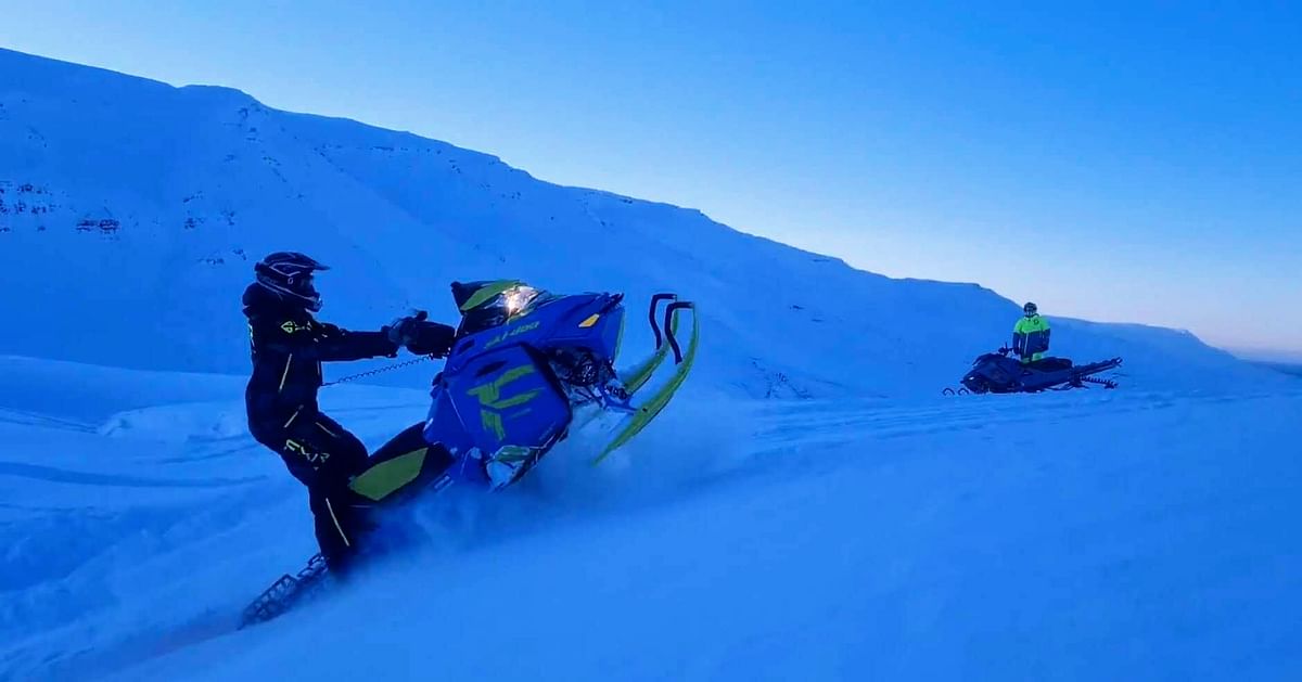 Two people riding snowmobiles on a snowy mountain under a clear blue sky.