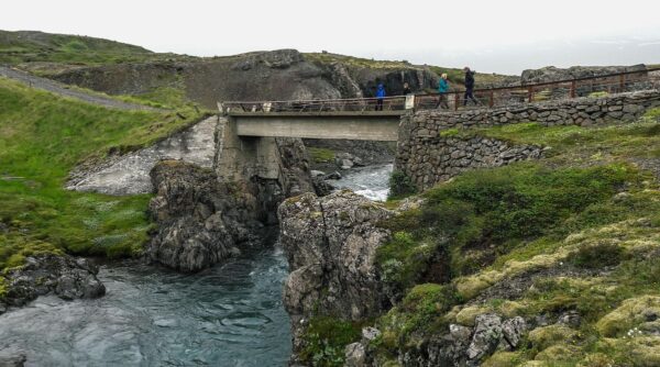 Bridge over rocky river surrounded by lush green hills, with people walking and enjoying the scenic landscape.