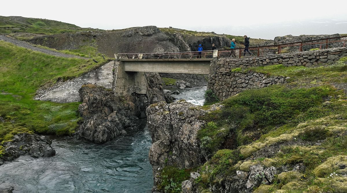 Bridge over rocky river surrounded by lush green hills, with people walking and enjoying the scenic landscape.