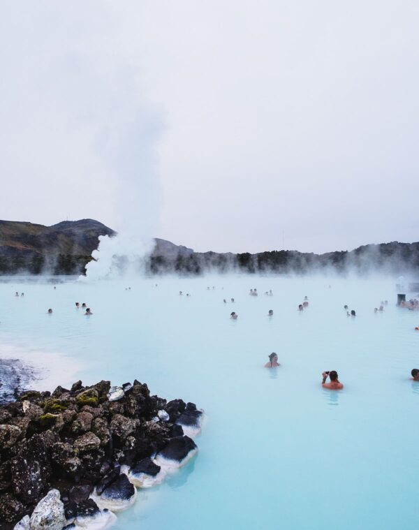 People enjoying the misty waters of the Blue Lagoon, Iceland, with steam rising and rocky edges visible.