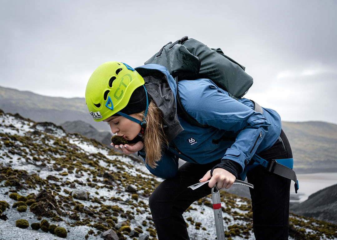 Woman wearing helmet and jacket kisses rock on a snowy hillside, holding an ice axe, with mountains in the background.