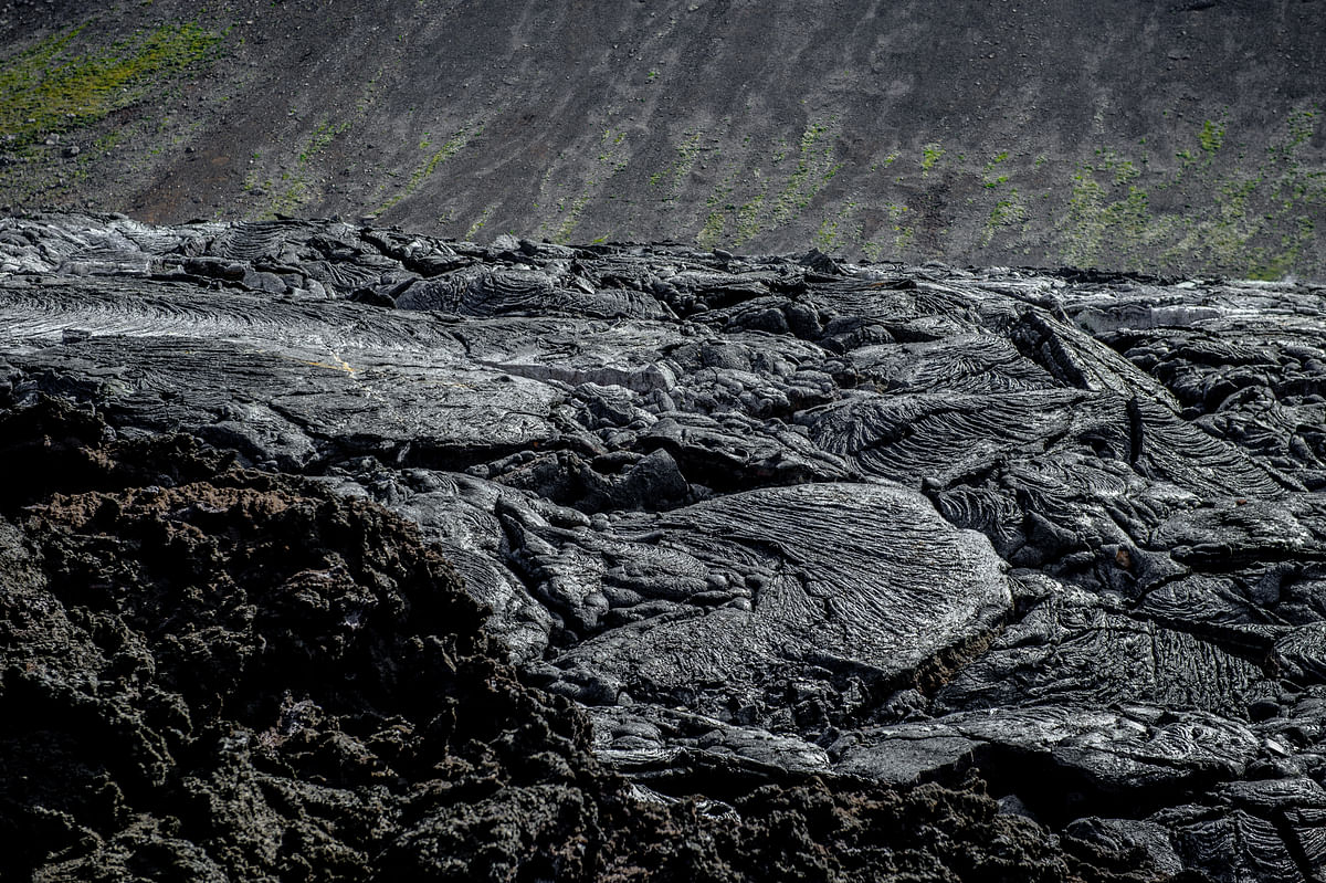 Textured volcanic lava flow with rugged, wavy surface patterns and partial grassy background.