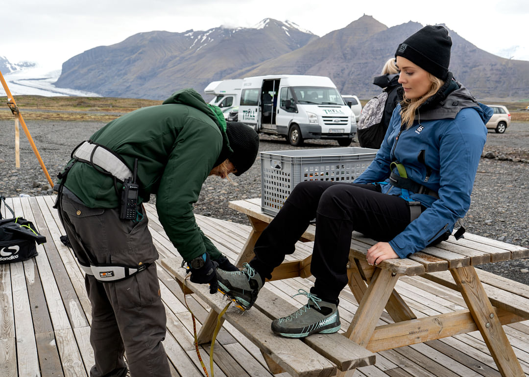 Guide helps woman prepare for glacier hike with crampons, mountains and vans in the background.