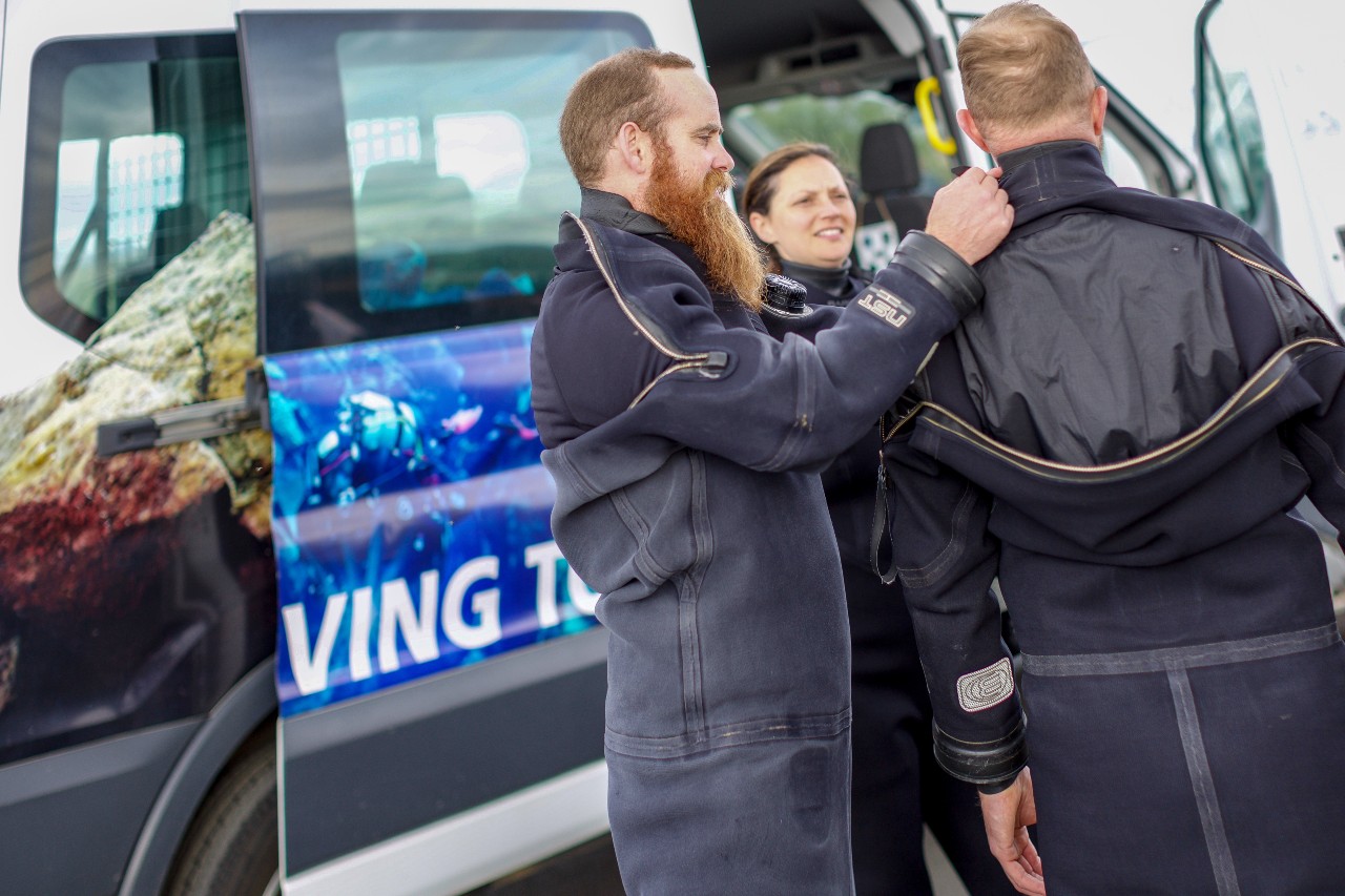 Divers preparing gear next to a diving tour van, ready for an underwater adventure in drysuits.