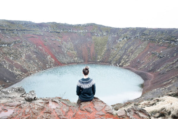 Person sitting at the edge of Kerid Crater, Iceland, overlooking the turquoise volcanic lake and colorful rocky slopes.