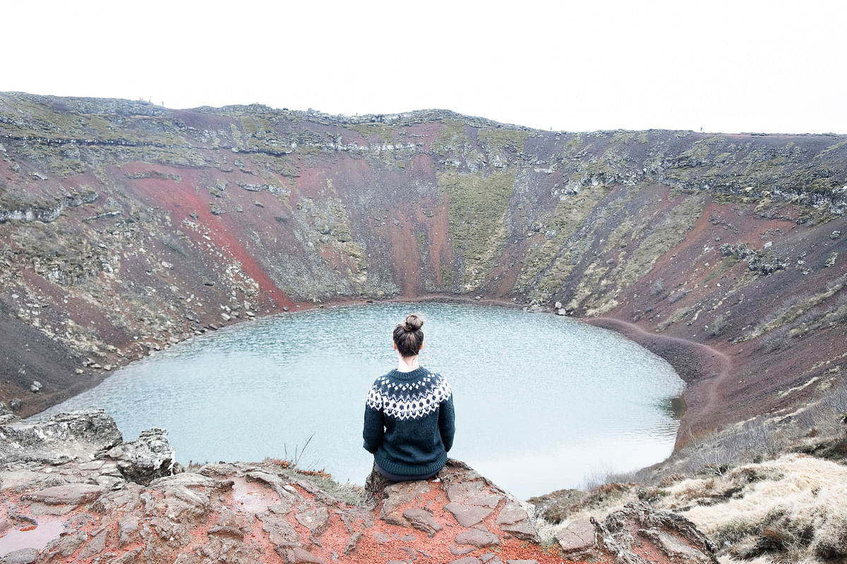 Person sitting at the edge of Kerid Crater, Iceland, overlooking the turquoise volcanic lake and colorful rocky slopes.