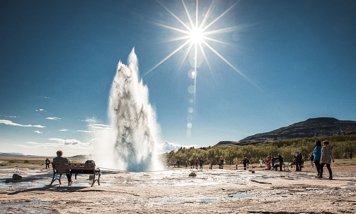 Geyser erupts under clear blue sky, tourists watch the spectacle with sun shining bright overhead in a scenic landscape.