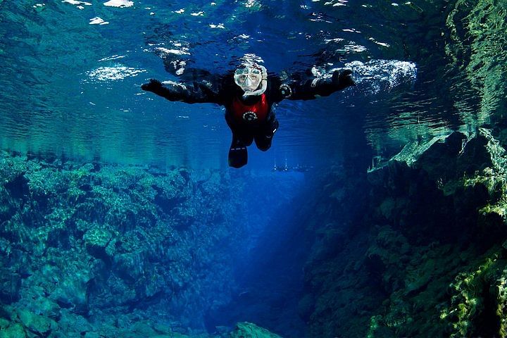 Person snorkeling in clear blue water over a rocky underwater landscape, wearing a wetsuit and snorkeling mask.