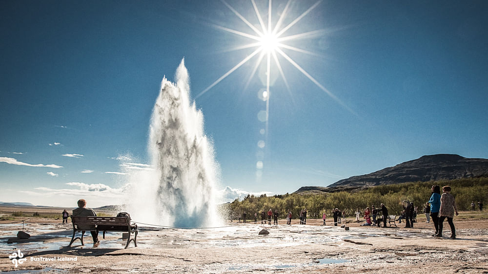 People watching a geyser erupt under clear blue skies and bright sun in a natural setting.