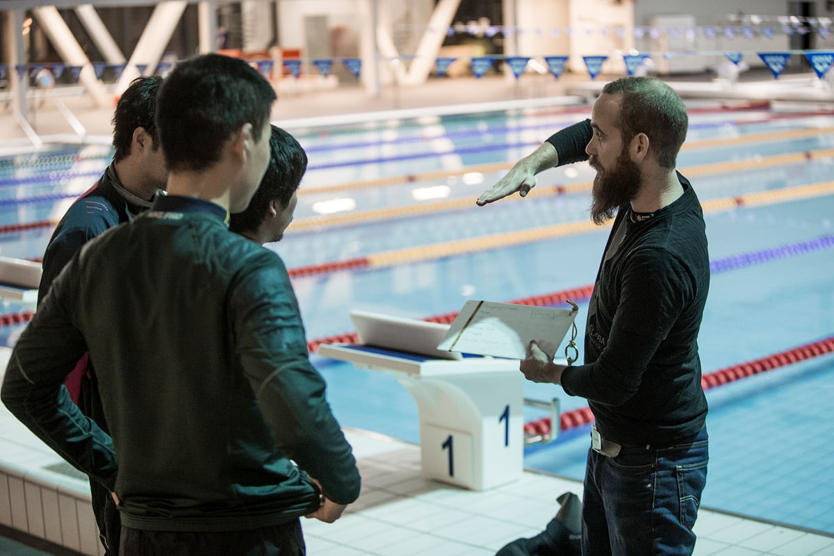 Swim coach giving instructions to swimmers poolside, holding clipboard, swim team training session.