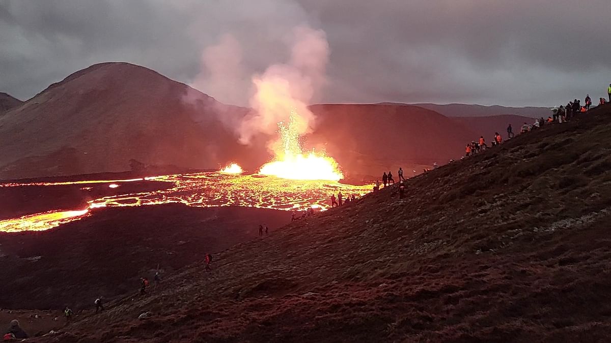 Lava flows from erupting volcano as onlookers gather on hillside, under a cloudy sky.