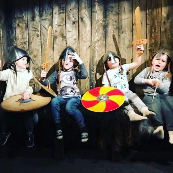 Four children dressed as Vikings with helmets, shields, and wooden swords, posing enthusiastically on a wooden bench.