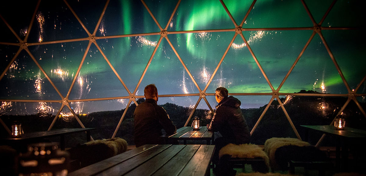 Two people watching Northern Lights from a cozy geodesic dome with candlelit tables.