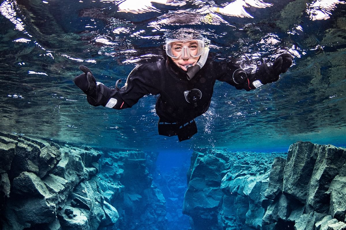 Diver in wetsuit snorkeling between tectonic plates in clear blue water, exploring underwater rock formations.