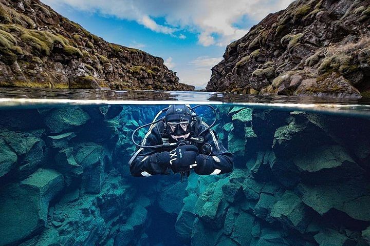 Diver explores underwater rift between tectonic plates in clear blue water, surrounded by rocky cliffs and bright sky.