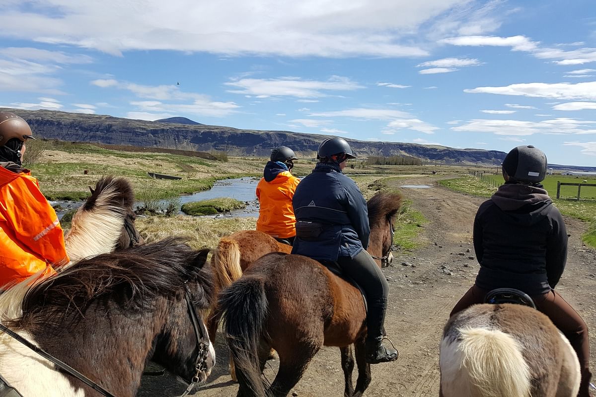 Horseback riders in gear explore a scenic, rural landscape with mountains and a river under a clear blue sky.