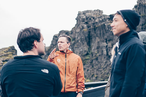 A group of people listening to a tour guide in an orange jacket with rocky cliffs in the background.