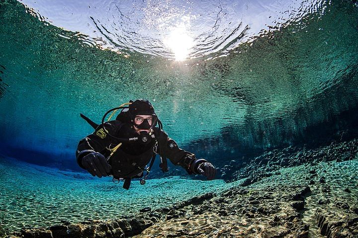 Scuba diver exploring a clear underwater cave with sunlight filtering through the surface, surrounded by rocky terrain.