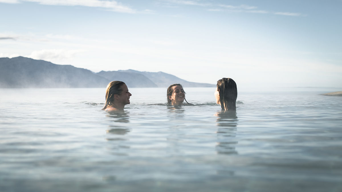 Three people enjoy swimming in a calm lake with mountains in the background on a clear day.