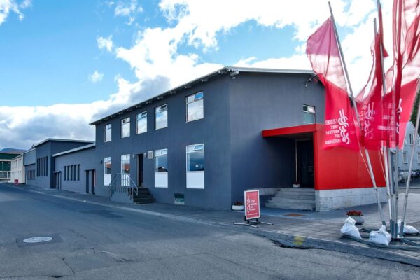 Modern gray building with red accents and flags, situated on a city street under a cloudy blue sky.