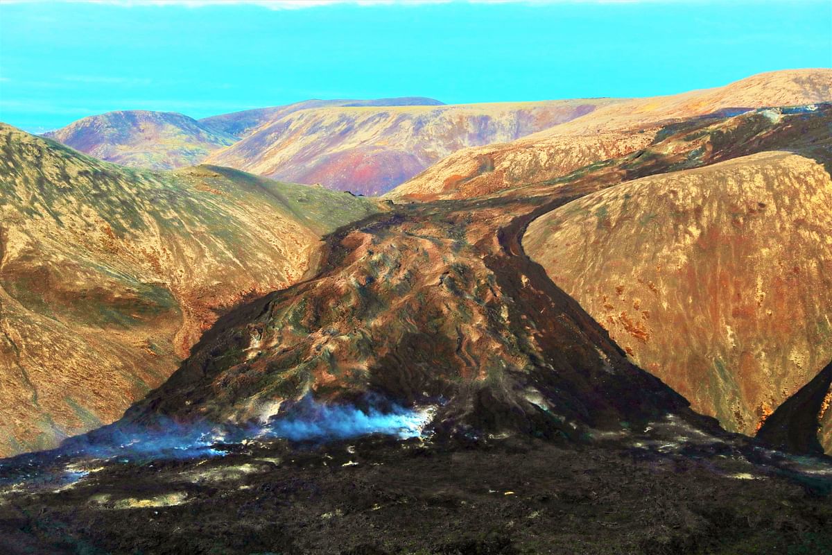 Aerial view of volcanic landscape with smoke rising, showcasing rugged terrain under a clear blue sky.