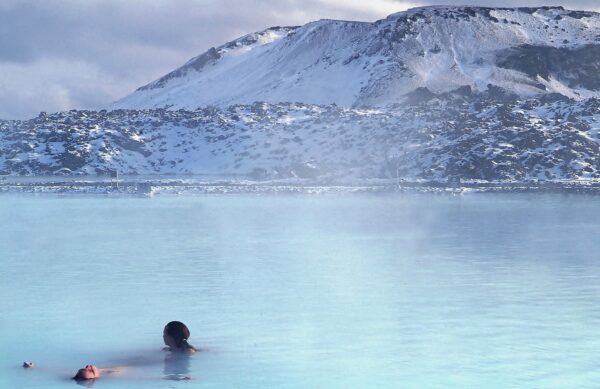 People relaxing in a steaming geothermal lagoon with snowy mountains in the background.