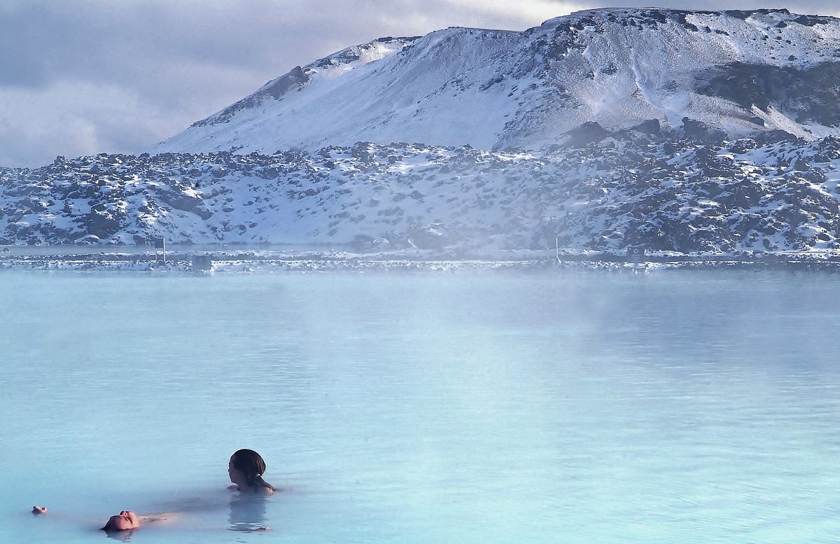 People relaxing in a steaming geothermal lagoon with snowy mountains in the background.