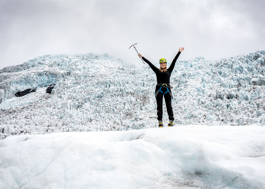 Person celebrating atop a snowy glacier, holding an ice axe with raised arms, showcasing adventure and achievement.