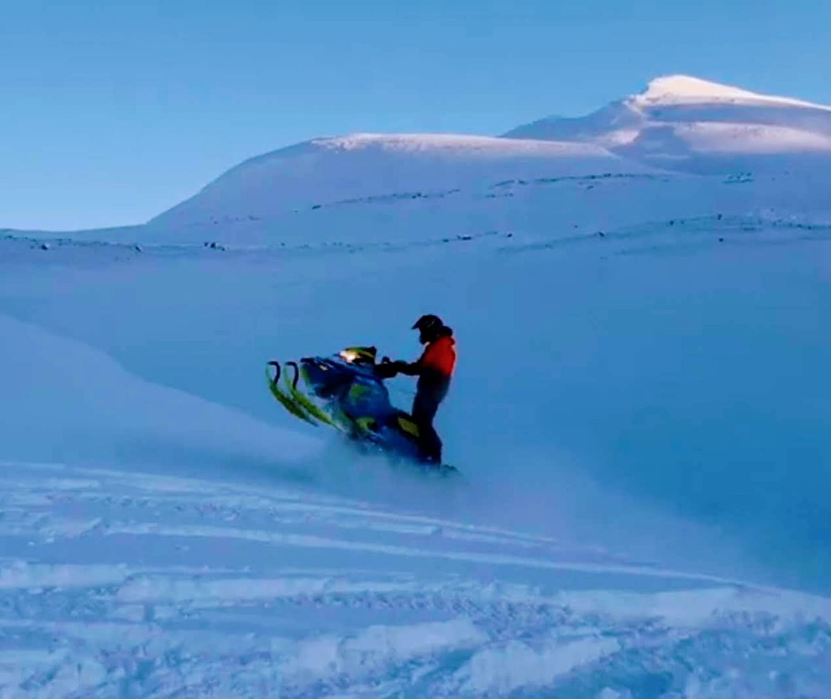 Snowmobiler rides over snowy landscape with mountains in the background under a clear blue sky.