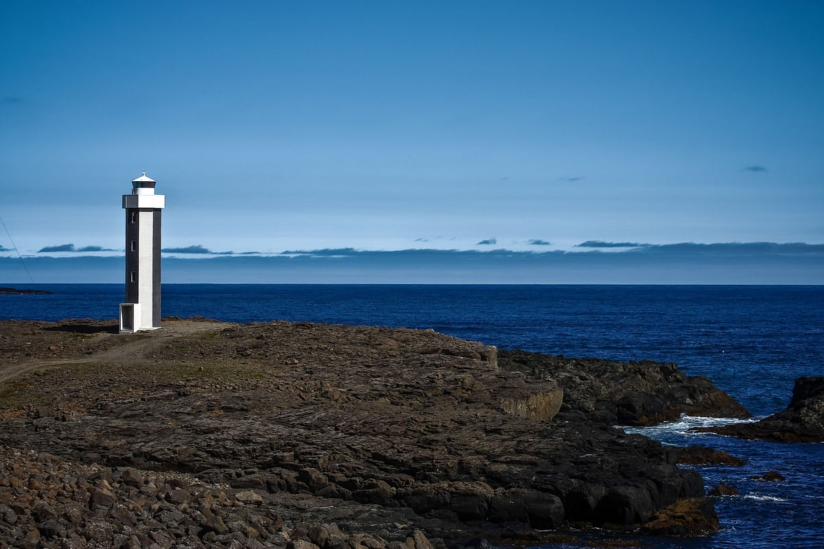 Tall lighthouse on rocky coastline against blue ocean and sky, ideal for navigation and nature themes.