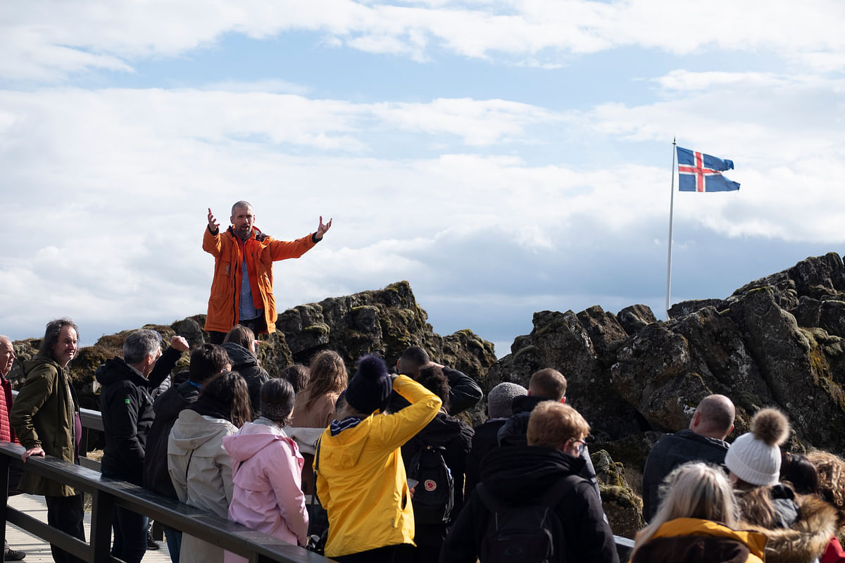 Tour guide addressing a crowd near rocks with Iceland flag in background under a cloudy sky.