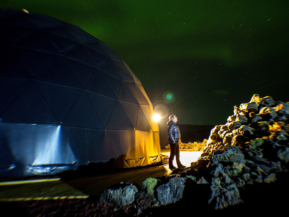 Person stargazing beside a geodesic dome under a green aurora-filled sky.