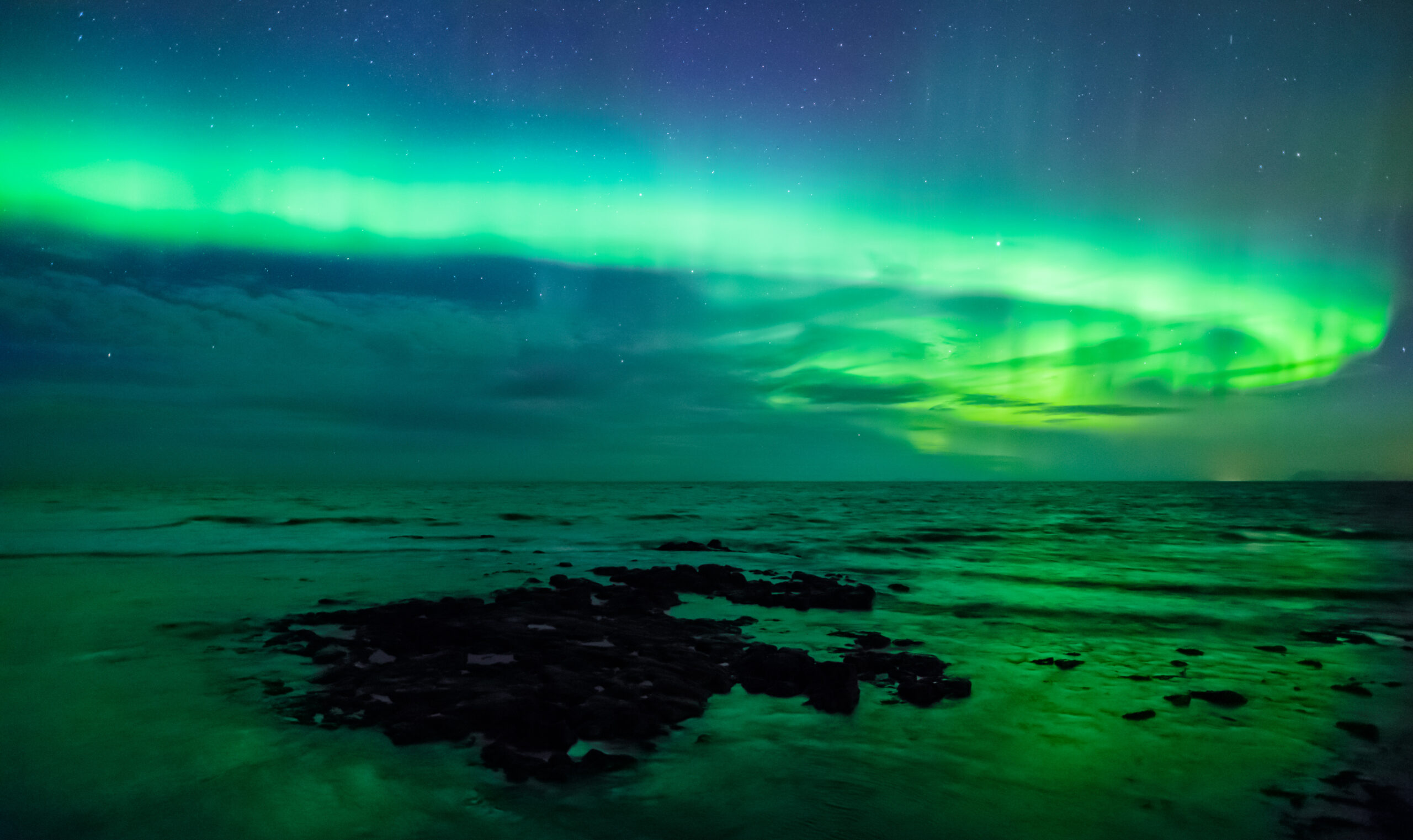 Aurora Borealis over the ocean, vibrant green and blue lights illuminating the night sky and reflecting on water.