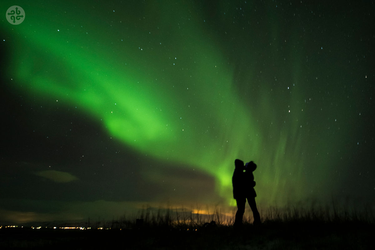 Silhouetted couple embraces under vibrant green Northern Lights in a starry night sky.