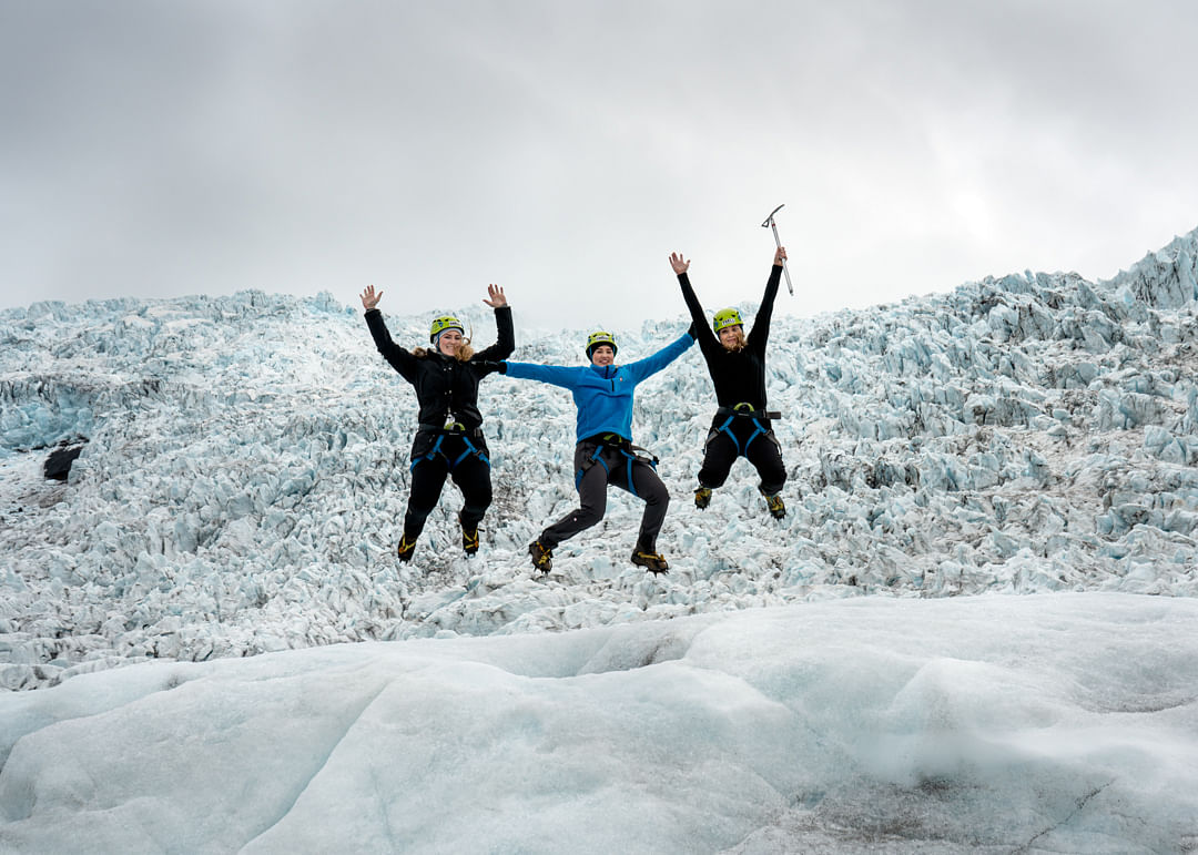 Three people jumping joyfully on a glacier, wearing climbing gear and helmets under a cloudy sky.