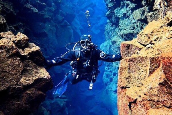 Scuba diver swims through underwater rock formation in clear blue waters.
