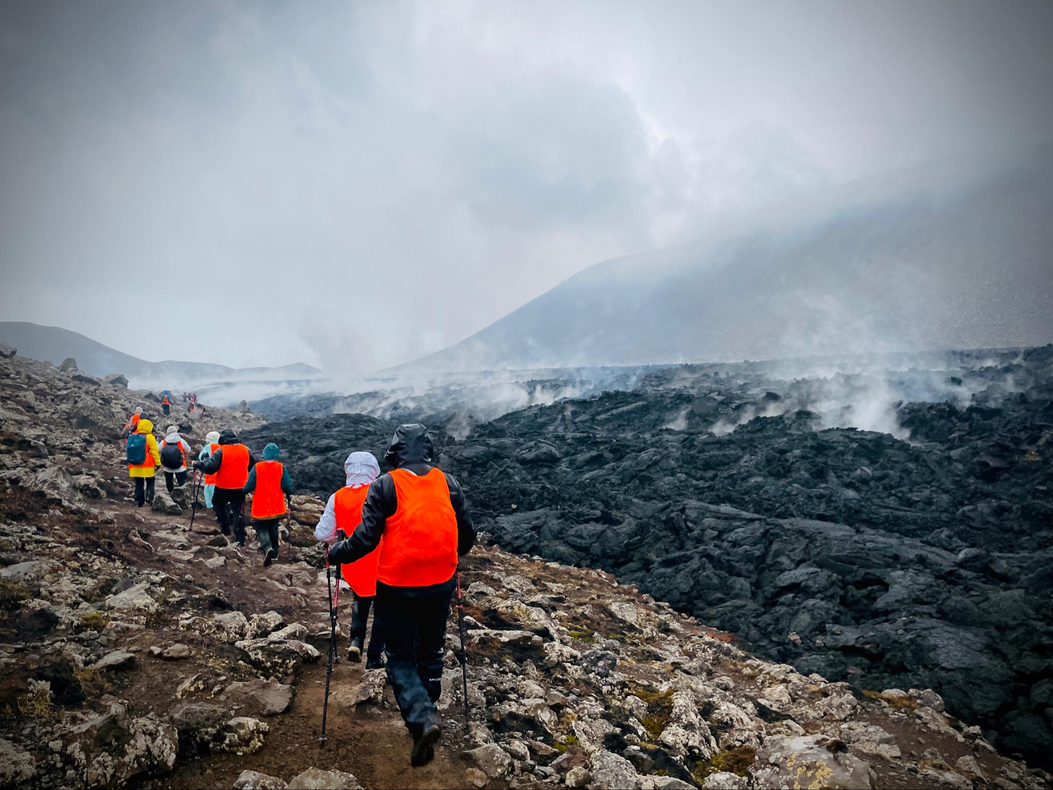 Group of hikers in orange vests walking near steaming volcanic lava field under cloudy skies.