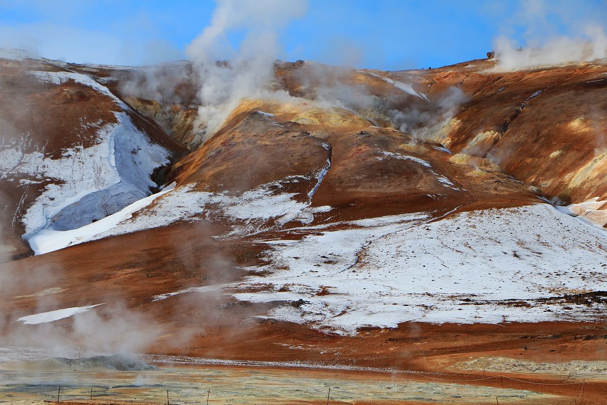 Steaming geothermal hills with snow patches in a rugged landscape under a clear blue sky.