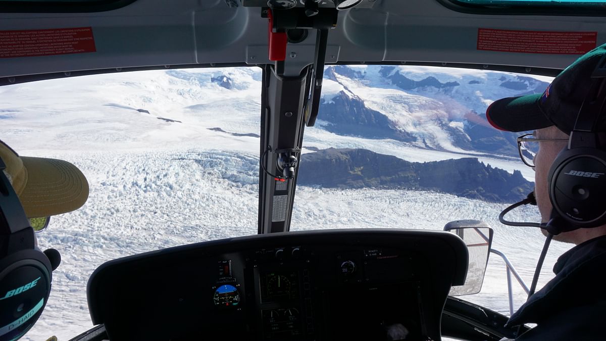 Inside a helicopter cockpit flying over a vast snowy glacier with rocky mountains in the background.