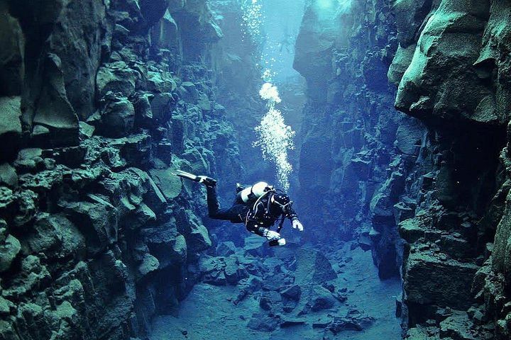 Scuba diver exploring the deep underwater crevices at Silfra fissure, Iceland, amidst rugged rocks and clear blue waters.