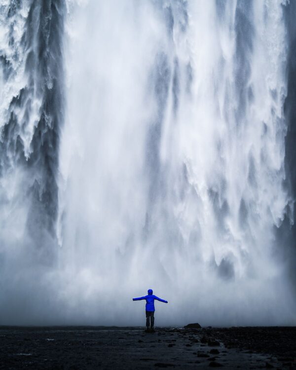 Person in a blue jacket stands with arms outstretched in front of a large, cascading waterfall.