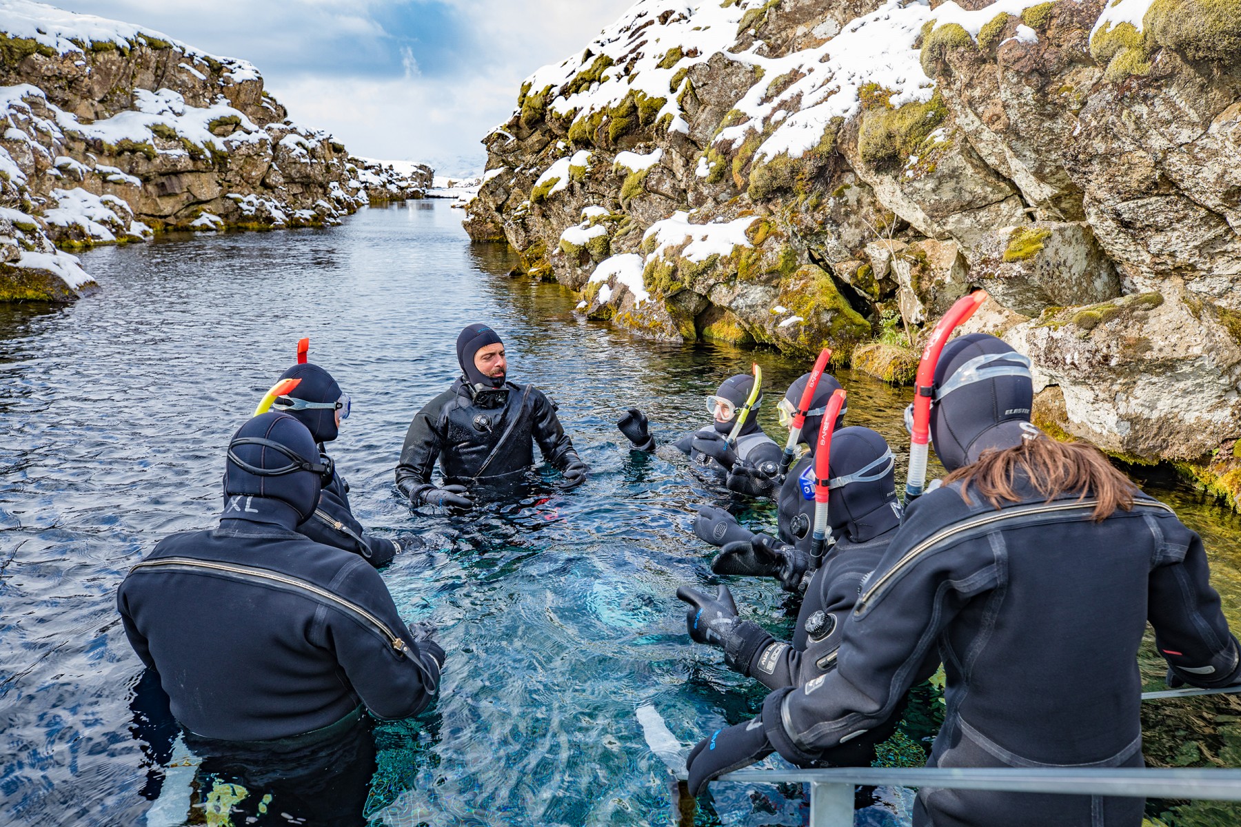 Snorkelers in wetsuits explore a snowy rocky waterway, guided by an instructor in clear water.
