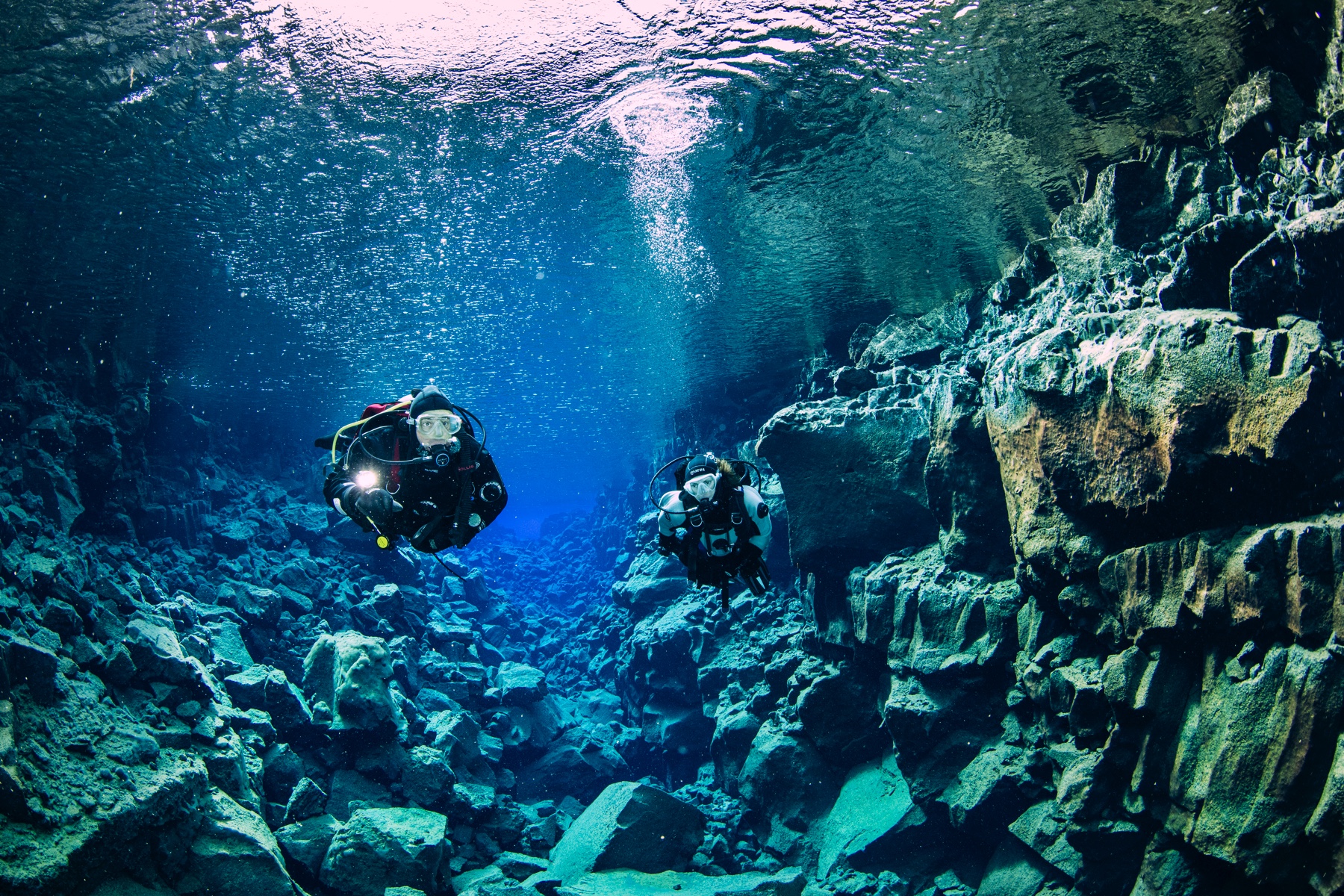 Scuba divers explore underwater volcanic rock formations in clear, blue water.