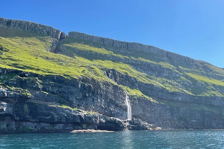 Cliffside waterfall cascading into tranquil blue waters under a clear sky, surrounded by lush green hills.