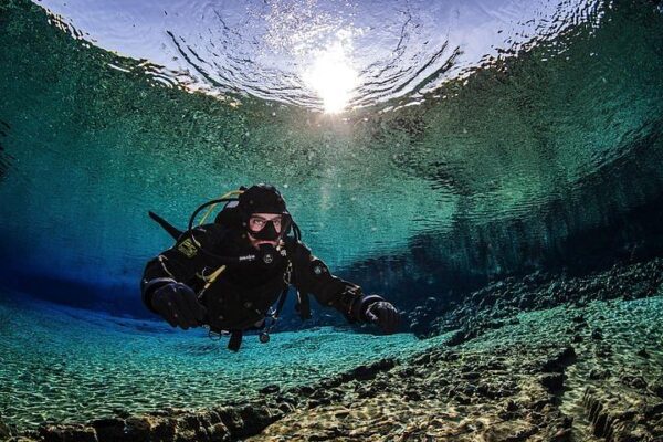 Scuba diver exploring crystal-clear underwater landscape with sunlight above, rocks and aquatic scenery.