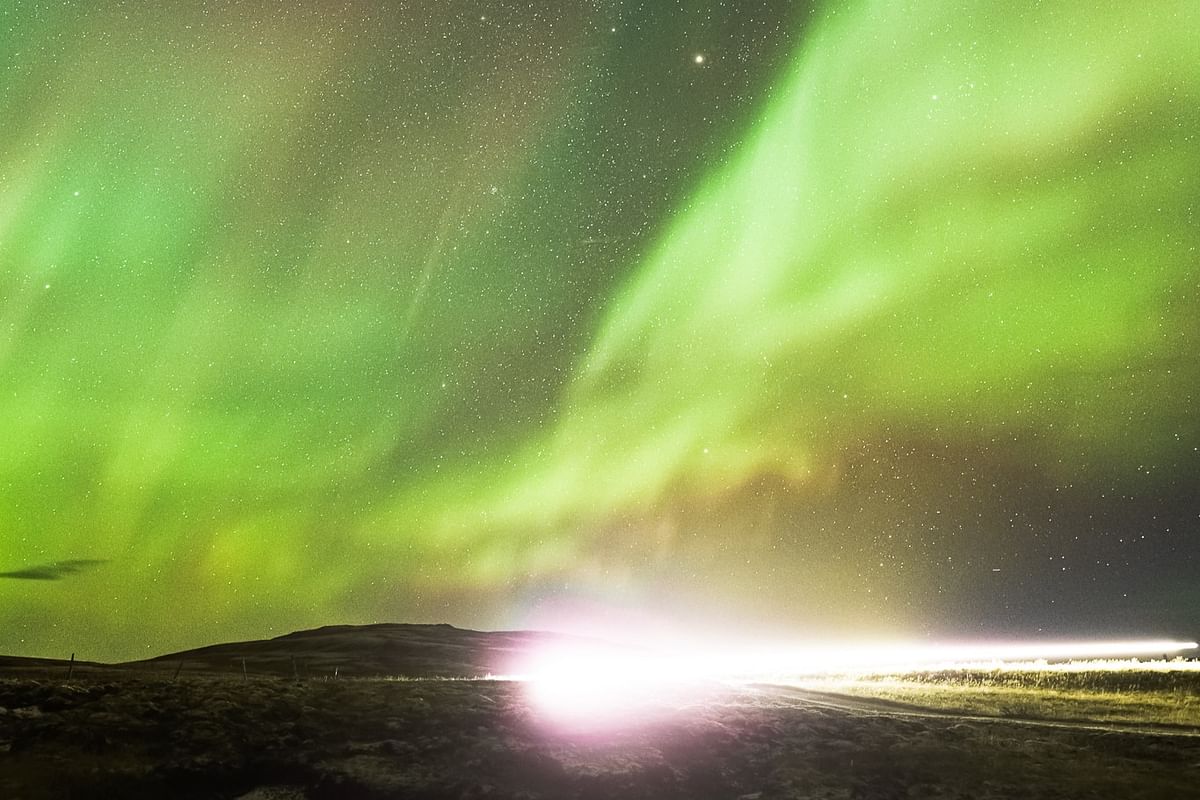 Vibrant green aurora borealis dancing over a dark landscape with bright lights illuminating the horizon.